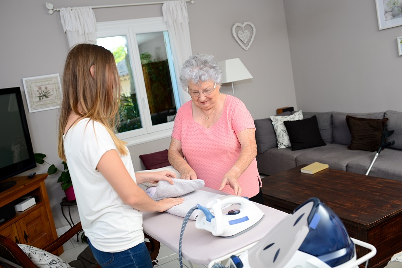 cheerful young girl ironing and helping elderly woman at home